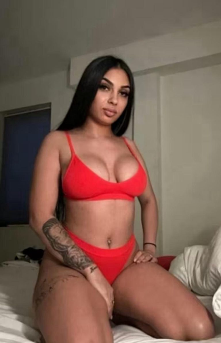 Woman in a red bikini seated on a bed, with long dark hair and a tattoo on her arm.